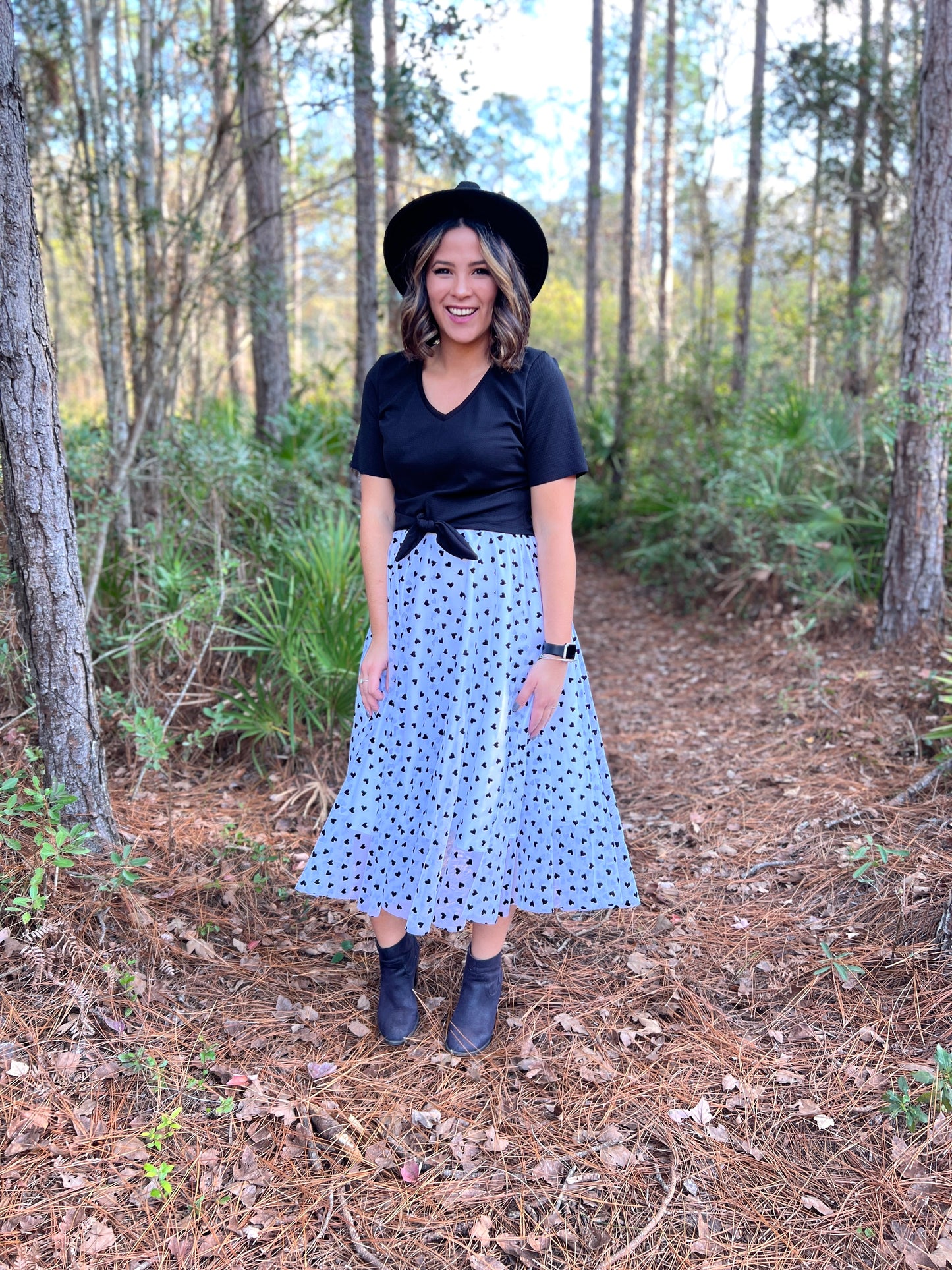 Woman standing on a trail in the woods wearing midi length white skirt with black dots & a black t-shirt