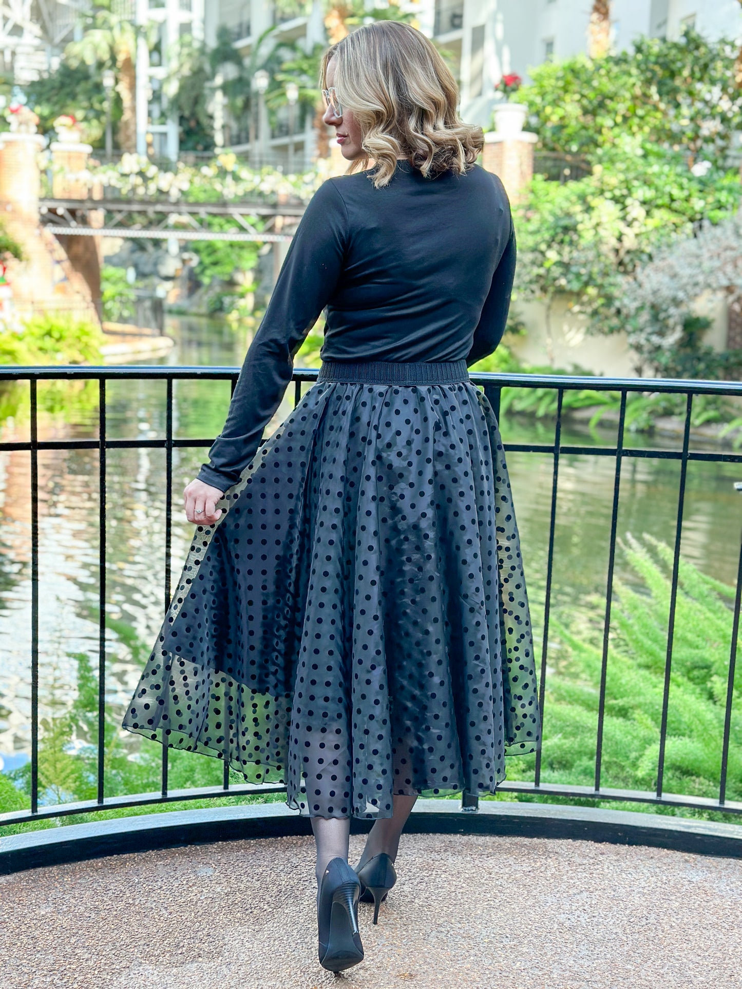 Woman standing on a bridge overlooking a garden river with a black enchated overlay skirt and a black top.