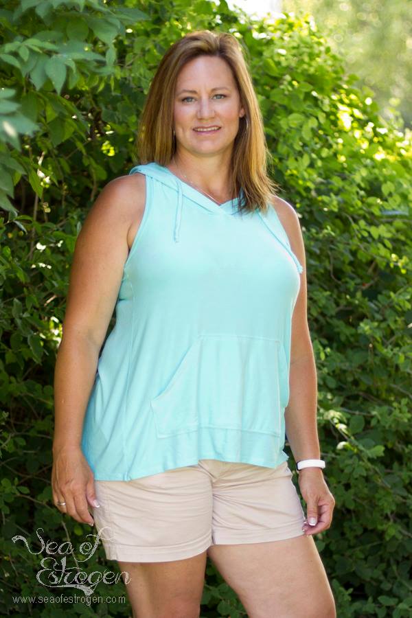 A woman standing in front of a hedge wearing a light teal Althea Tank Top with the hood down.