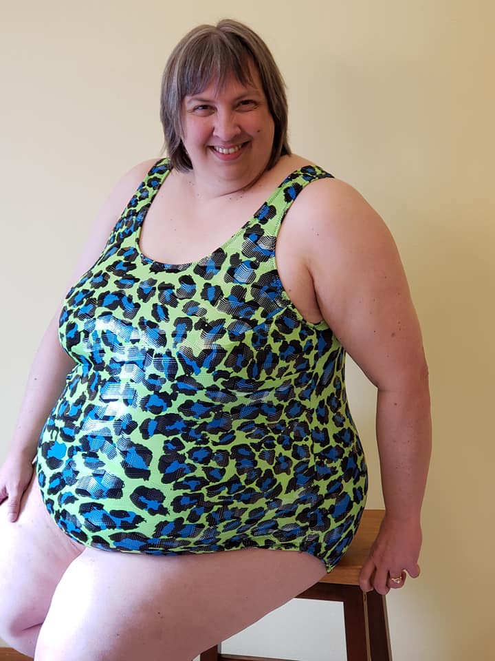 Plus size woman sitting on a stool wearing a green, blue, and black DIBY Club Amelia One-piece swimsuit with leopard print pattern.