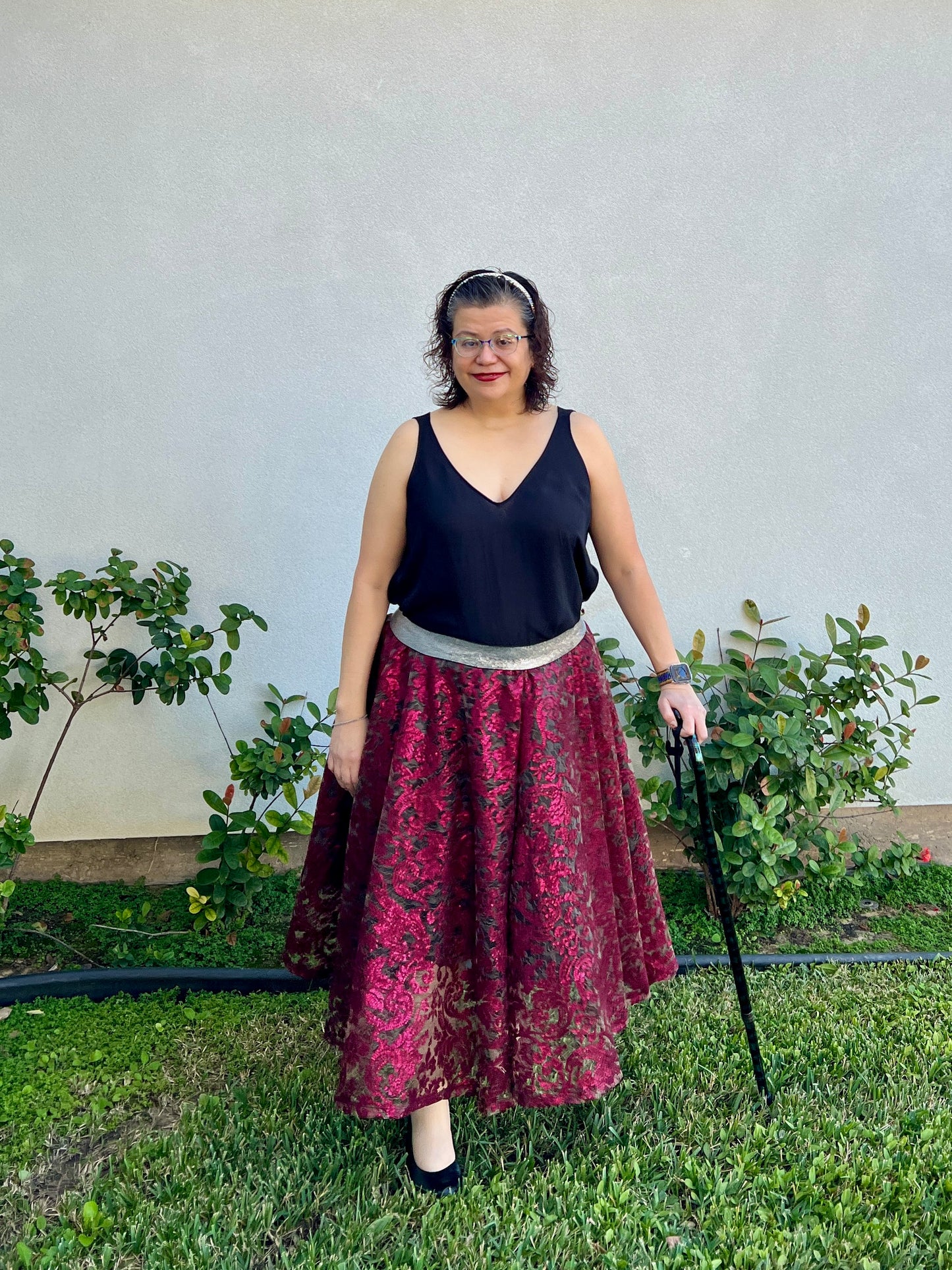 Woman standing with a cane next to a small garden wearing the enchanted overlay skirt with a dark red lace overlay and a black tank top.