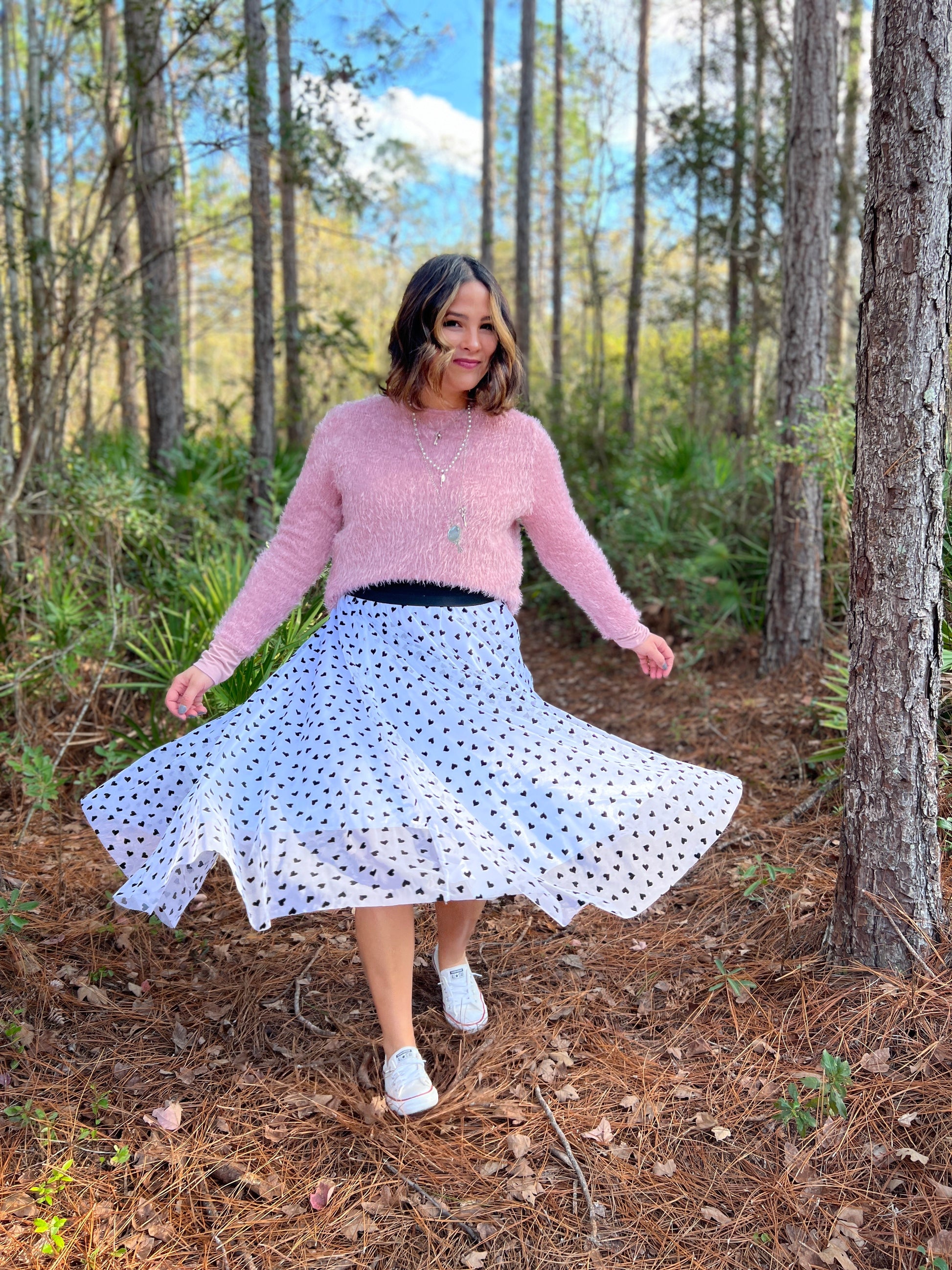Woman twirling her white and black enchanted overlay skirt wearing a pink sweater on a trail in the woods
