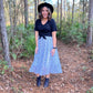 Woman standing on a trail in the woods wearing midi length white skirt with black dots & a black t-shirt
