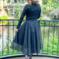 Woman standing on a bridge overlooking a garden river with a black enchated overlay skirt and a black top.