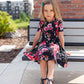 A young girl sits on a bench while wearing a floral themed, short-sleeved, knee length Samantha Gathered Dress. 